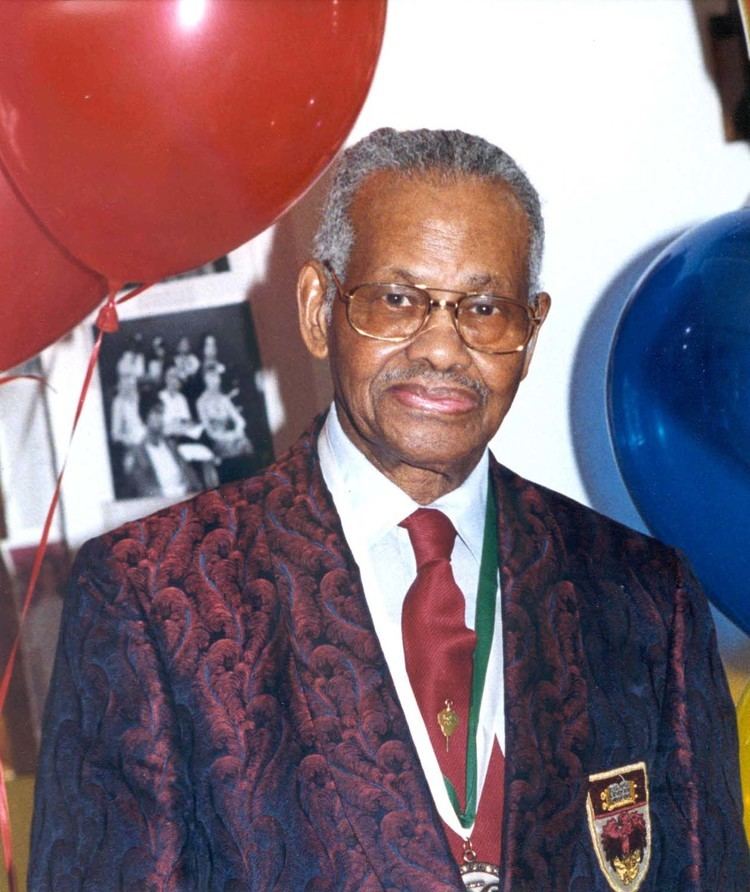 Leonidas Berry smiling with red and blue balloons in his background during the Rush Distinguished Alumnus Award in 1987, having gray hair, wearing eyeglasses, and a medal, a white polo shirt, a tie pin and a red necktie under a red coat with a logo