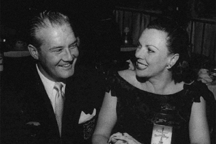 George Reeves smiling and wearing a suit and a tie while looking at Leonore Lemmon wearing a black dress.