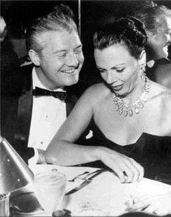 George Reeves smiling while looking at Leonore Lemmon wearing a necklace and a black dress.