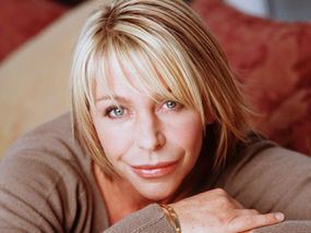 Leslie Ash smilies while sitting and wearing a brown long sleeve