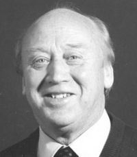 Leslie Norris with a smiling face, wearing a suit, a tie, and white long sleeves.