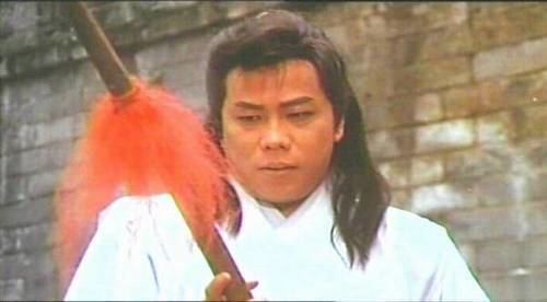 Leung Siu-lung with a serious face while looking at the spear with an orange decoration, with long hair, and wearing a white robe.