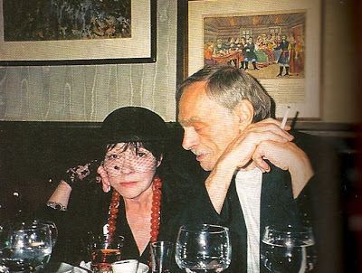 Lev Zbarsky looking at the woman beside him who is wearing black hat, black blouse and red necklace