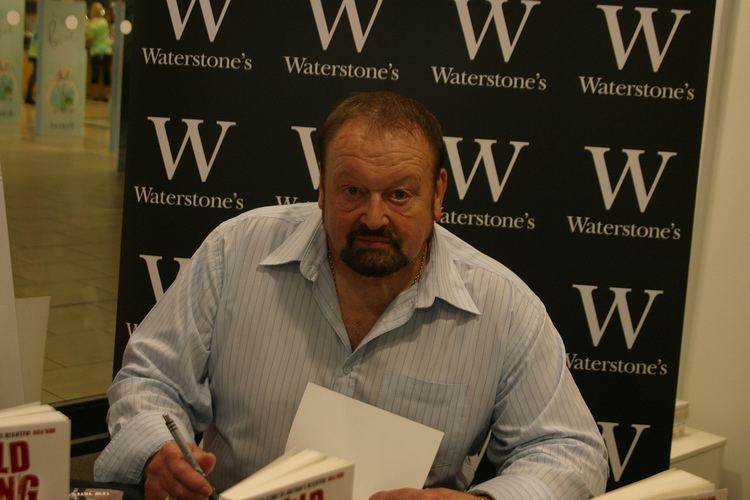 Lew Yates signing a book while wearing a light blue striped long sleeves