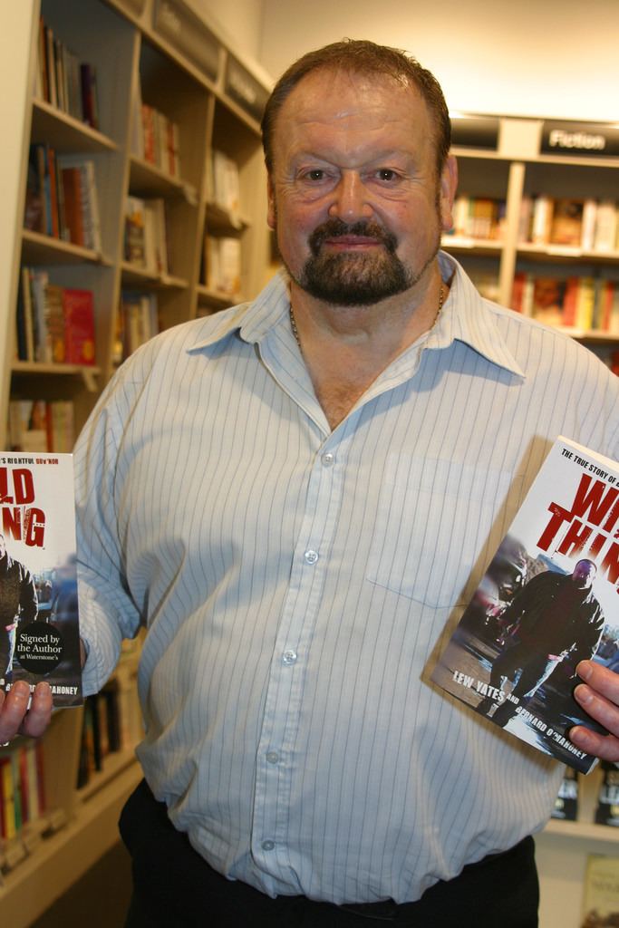 Lew Yates with a tight-lipped smile, mustache, and beard while holding two books and he is wearing a light blue striped long sleeves