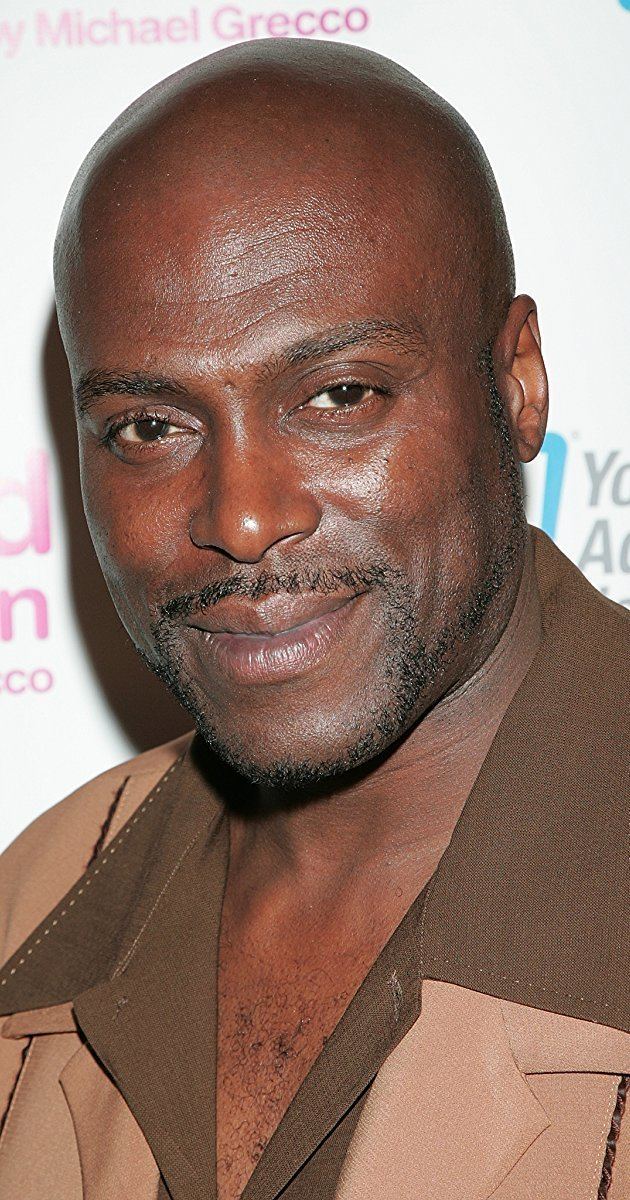 Lexington Steele smiling while wearing a beige coat and brown long sleeves
