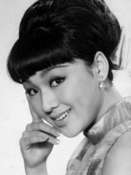 Li Ching smiling, with bangs and her hair tied up, and wearing a sleeveless blouse and earrings