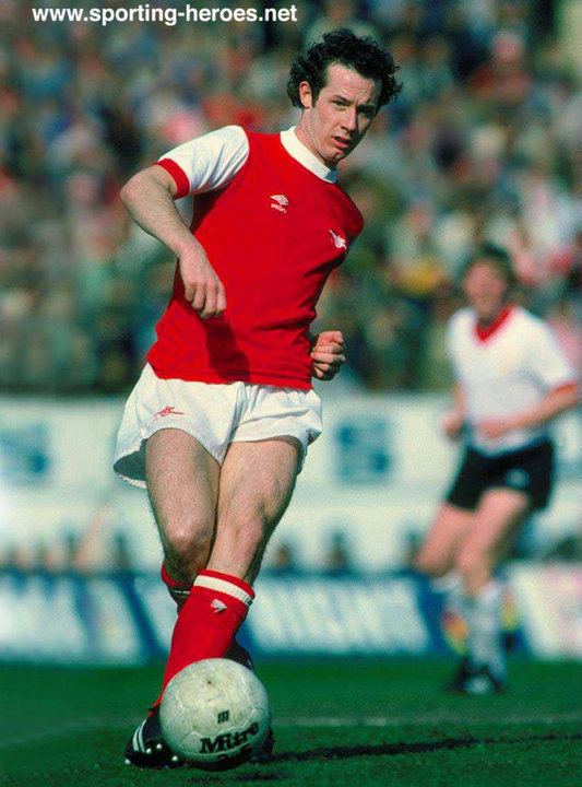 Liam Brady wearing a red and white shirt, white shorts, red socks, and black shoes while playing football.