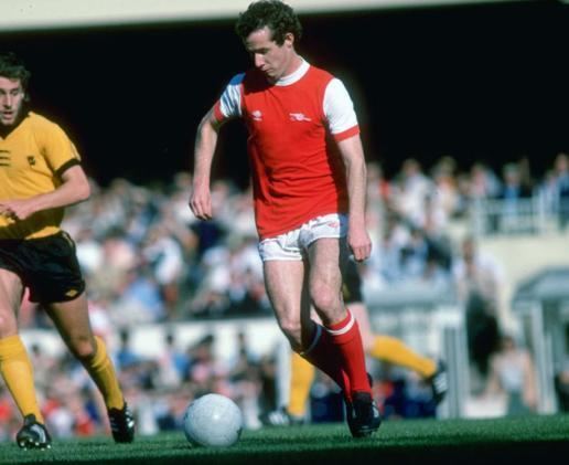Liam Brady wearing a red and white shirt, white shorts, red socks, and black shoes while playing football.