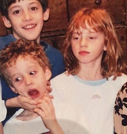 Lily Newmark with her two brothers. Lily's brother is holding the face of his younger brother while Lily is looking at something and wearing a white blouse