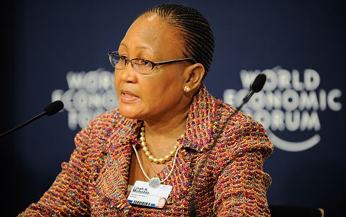 Linah Mohohlo wearing eyeglasses, earrings, a necklace, and a colorful dress with a microphone in front of her.