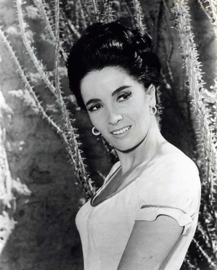 Linda Cristal smiling with a tied-up hairstyle and wearing a blouse and earrings