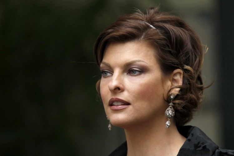 Linda Evangelista looking afar with short curly hair and her mouth is half-open while wearing earrings, a headband, and a black blouse