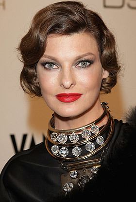 Linda Evangelista with a tight-lipped smile, gray eyes, and brown short wavy hair while wearing a necklace with gemstones and a black blouse