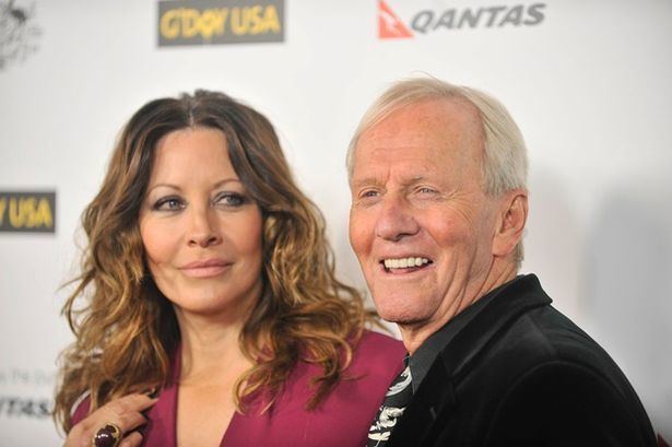 Linda Kozlowski with a tight-lipped smile and looking at something while Paul Hogan is smiling. Linda with wavy blonde hair, wearing a black ring and a maroon top while Paul with white hair, wearing a black coat over black long sleeves, and a white and black necktie.