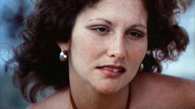 Linda Lovelace is looking at something with a serious face, with curly short hair, wearing earrings and a red halter top.