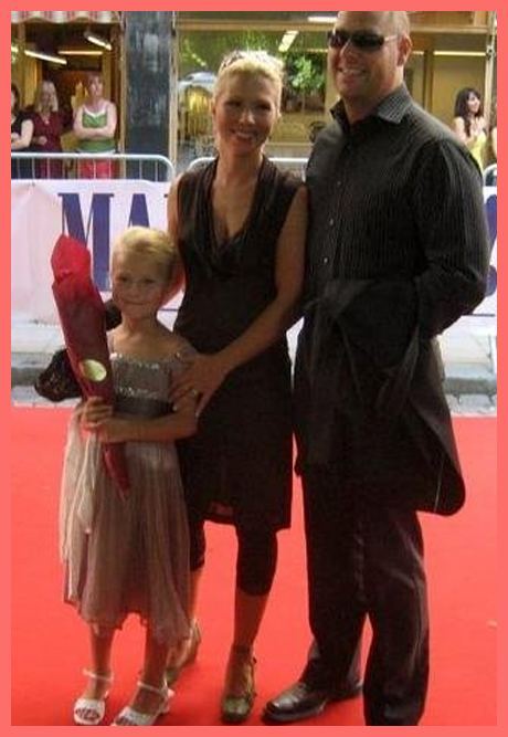 Linda Ulvaeus smiling while wearing a brown dress with her partner and daughter