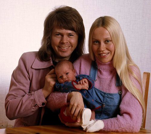 Agnetha Faltskog smiling and wearing a pink sweatshirt with Bjorn Ulvaeus while carrying a baby