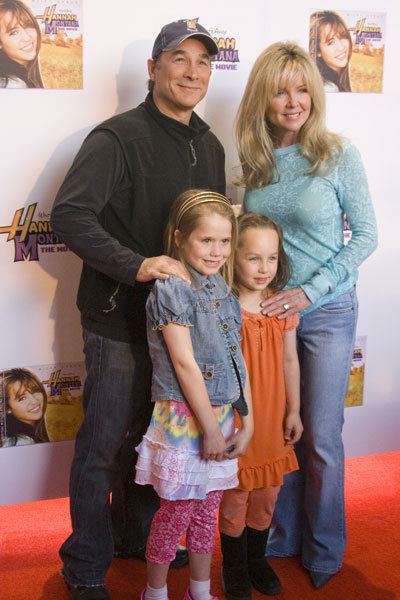 In the Hannah Montana Event, At the back from left, Clint Black (left) is smiling, standing next to Lisa Hartman Black, has brown hair, wearing a blue cap, black jacket, denim pants and black shoes, at the right, Lisa Hartman Black (right) is smiling, posing with her right side facing the wall, standing in front of Hannah Montana backdrop, has blonde hair, gray eyes, wearing silver ring, cyan long sleeve, denim pants and black shoes, In front, Chelsea Bain (left) is smiling, standing, hands down, has brown hair, wearing a gold headband, white colorful dress under a denim jacket and pink knee length short and white socks, Lily Pearl Black (right) is smiling, standing hands on her side, has brown hair, wearing a orange dress, and orange pants with black boots.