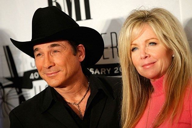 On the left, Clint Black is smiling, standing, has black hair wearing a cowboy hat, silver necklace, black coat, At the right Lisa Hartman Black is smiling, standing in front of a white backdrop, has blonde hair, gray eyes, wearing a silver necklace, silver earrings and pink turtle neck shirt.