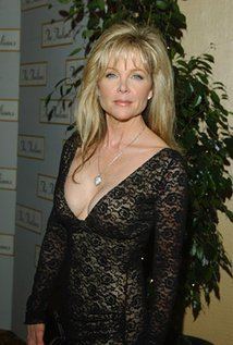 Lisa Hartman Black is serious, standing in front of a green backdrop wall, and tall plant, has blonde hair, gray eyes, wearing a silver necklace, earrings and a cleavage showing black long sleeve dress.