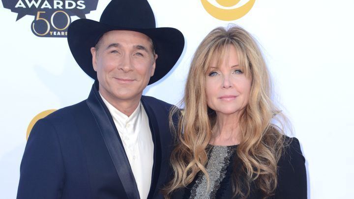 In ACM AWARDS Event, On the left, Clint Black is smiling, standing, has brown hair wearing a cowboy hat, white top and black coat, At the right Lisa Hartman Black is serious, standing in front of a white ACM awards backdrop has blonde hair, gray eyes, wearing a black with silver dress.