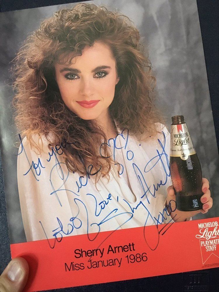 Sherry Arnett, a Playboy Playmates of 1986, smiling while holding a Michelob beer and wearing a white blouse with an autograph over it