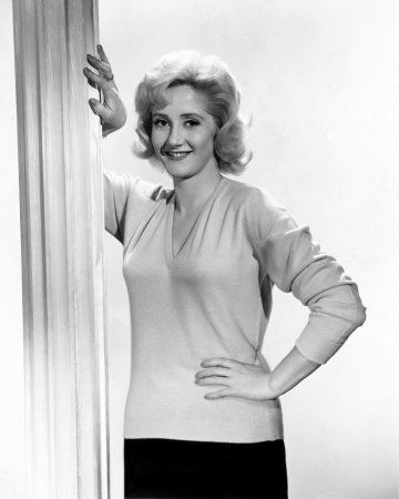 Liz Fraser smiling, with curly hair, wearing a knitted blouse and black skirt.