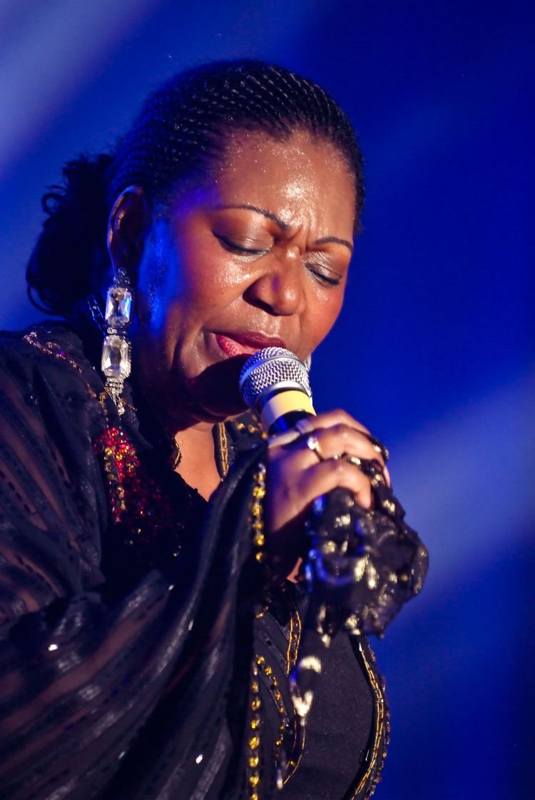 Liz Mitchell singing with her eyes closed and braided hair while wearing a black dress and dangling earrings