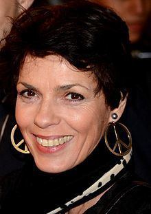 Élizabeth Bourgine smiling with short hair and wearing a black coat, black and white scarf, and gold round earrings