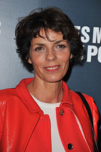 Élizabeth Bourgine smiling with short hair, wearing a white shirt and orange coat, and carrying a black bag