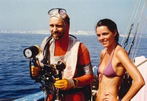 Élizabeth Bourgine smiling and standing beside a man on a yacht with hands on the waist and wearing a pink bikini