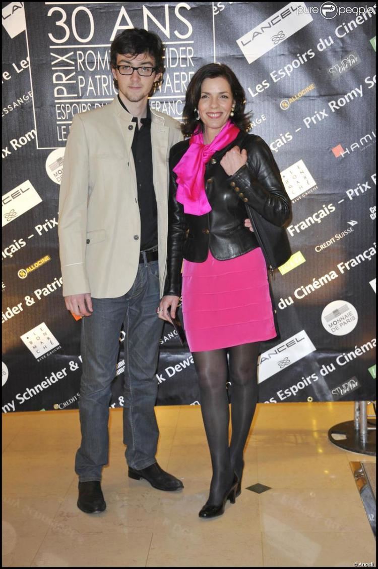 Jules Bourgine smiling and wearing a black shirt under a brown coat, gray pants, belt, and black shoes during the Romy Schneider And Patrick Dewaere Awards 2011 at Le Bon Marche in Paris with Elisabeth Bourgine wearing a pink dress under a black leather coat, pink scarf, black stockings, black heels, and pearl earrings