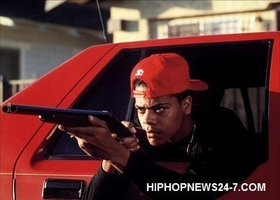 Scene from Boyz n the Hood movie, featuring Lloyd Avery II wearing a red cap and holding a gun inside a red car.