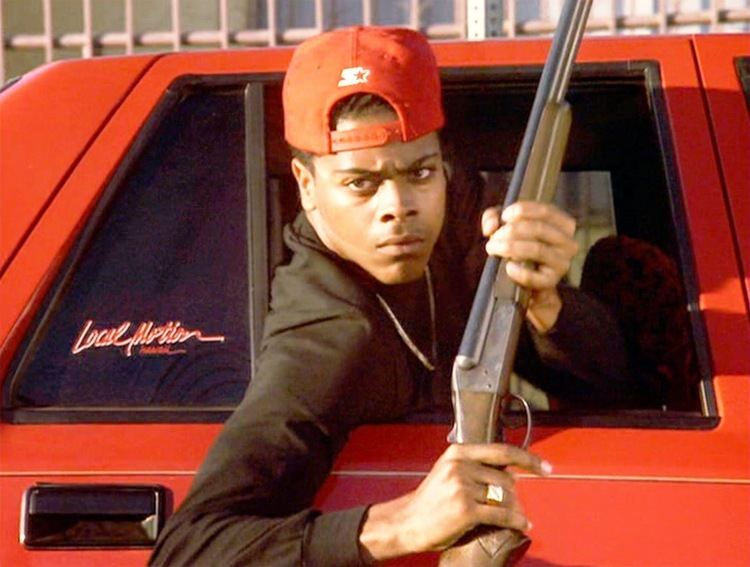 Scene from Boyz n the Hood movie, featuring Lloyd Avery II wearing a red cap, a necklace, a ring, and holding a gun inside a red car.