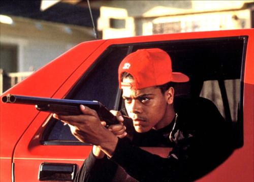 Scene from Boyz n the Hood movie, featuring Lloyd Avery II wearing a red cap and holding a gun inside a red car.
