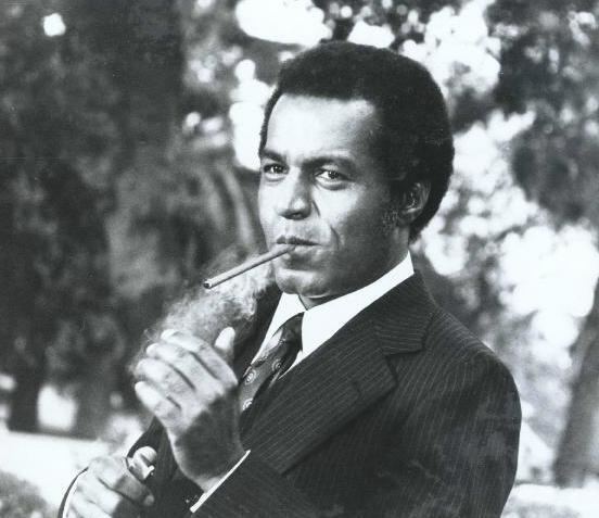 Lloyd Haynes smoking while wearing a striped coat, white long sleeves, and necktie