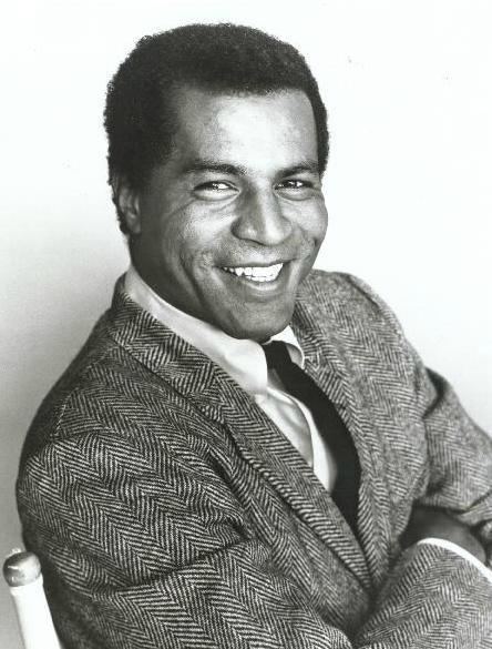 Lloyd Haynes smiling while wearing a patterned coat, long sleeves, and necktie