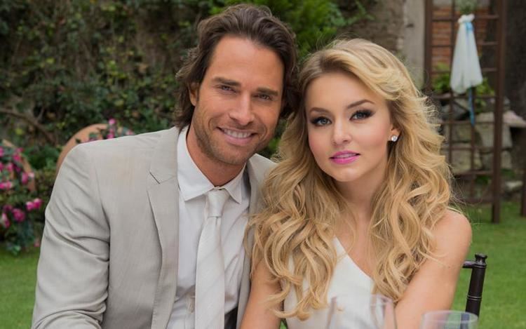 Sebastián Rulli and Angelique Boyer are smiling while sitting on a chair with a garden in their background. Sebastián is wearing a white long sleeve under a white necktie and gray coat. Angelique is wearing earrings and a white one-sided shoulder top