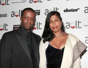 Lolita Chakrabarti smiling and wearing a cream and gray blazer and black blouse while Adrian Lester wearing a black coat and long sleeves