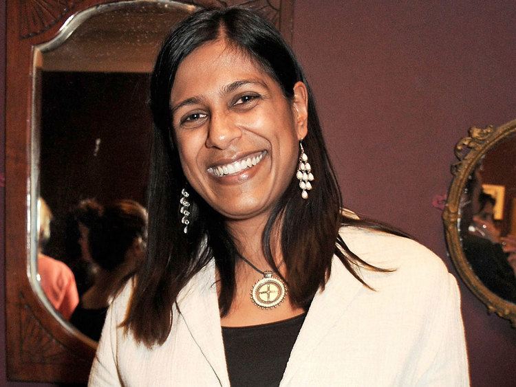 Lolita Chakrabarti smiling while wearing a cream coat, black inner blouse, necklace, and earrings