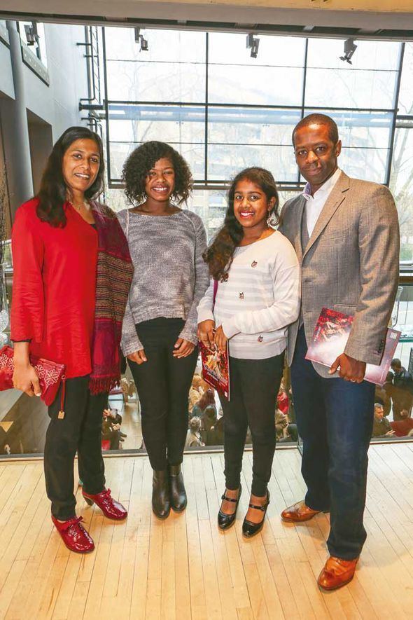 Lolita Chakrabarti smiling with her husband and two daughters while wearing a red long sleeves blouse, black pants, and red shoes