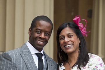Lolita Chakrabarti smiling with Adrian Lester while wearing a white floral blouse, earrings, and pink ribbon on her hair