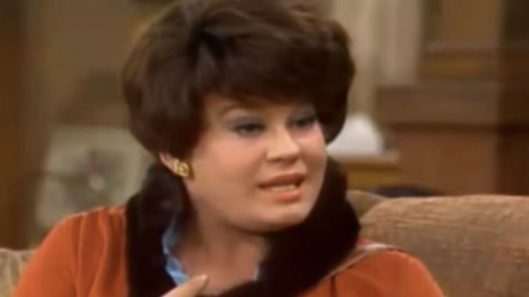 Lori Shannon talking to someone while wearing an orange blouse and black scarf in a scene from the 1971 tv series, All in the Family-Archie the Hero