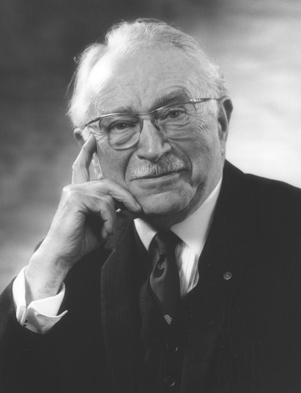Ludwig Guttmann wearing eyeglasses, a black suit, and a tie while holding his face.