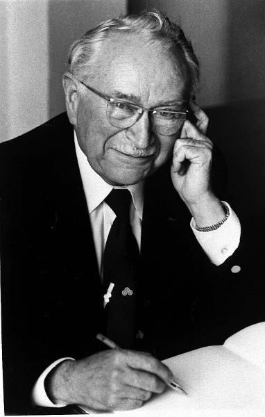 Ludwig Guttmann wearing eyeglasses, a black suit, and a tie while holding his face and with a pen on his hand.