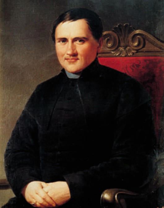 Luigi Caburlotto posing in a portrait holding his hands and sitting in a red chair while wearing black cassock (clergy garments).