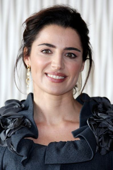 Luisa Ranieri smiling with tied-up hair while wearing a dark blue blouse with a ruffle neckline and earrings