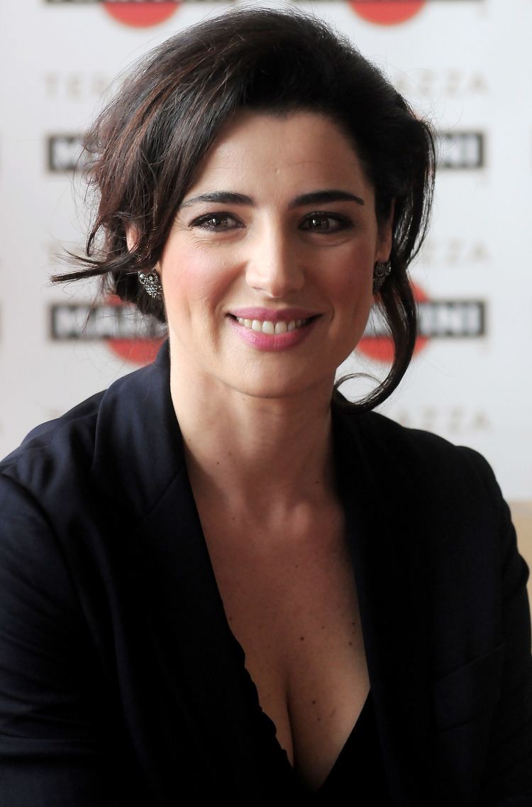 Luisa Ranieri smiling while looking at something with black messy hair and wearing earrings and a black blouse under a black coat that exposes her cleavage