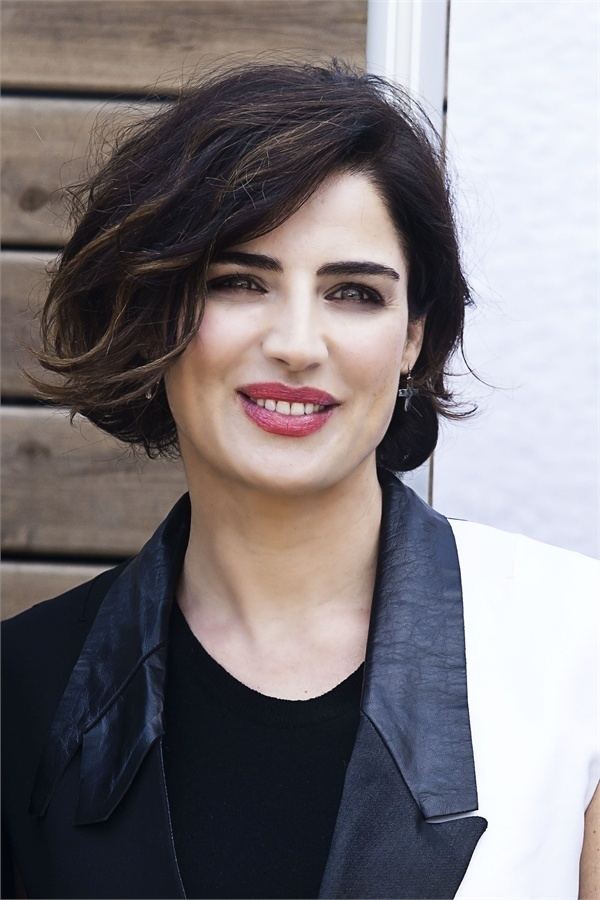 Luisa Ranieri smiling with short wavy hair while wearing earrings and a black blouse under a black and white coat